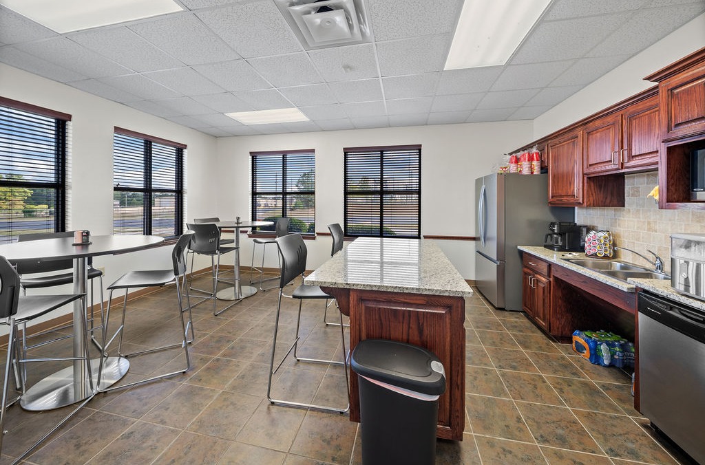 Office break room with a granite-topped island, bar seating, kitchen cabinets and a stainless refrigerator.