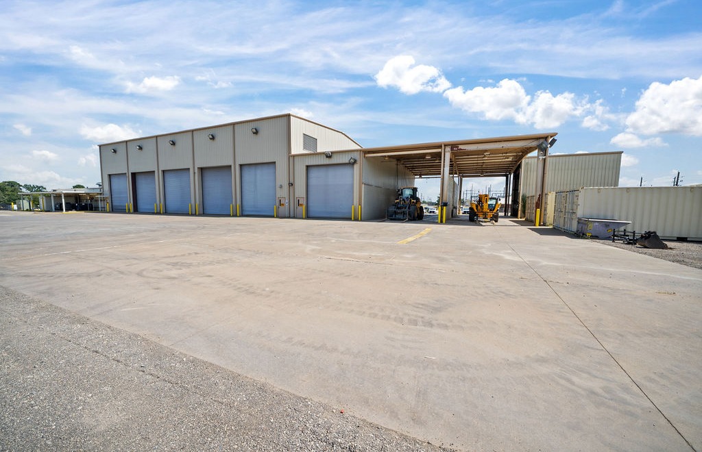 Metal industrial shop with multiple roll-up overhead doors beside a covered equipment bay and paved yard.
