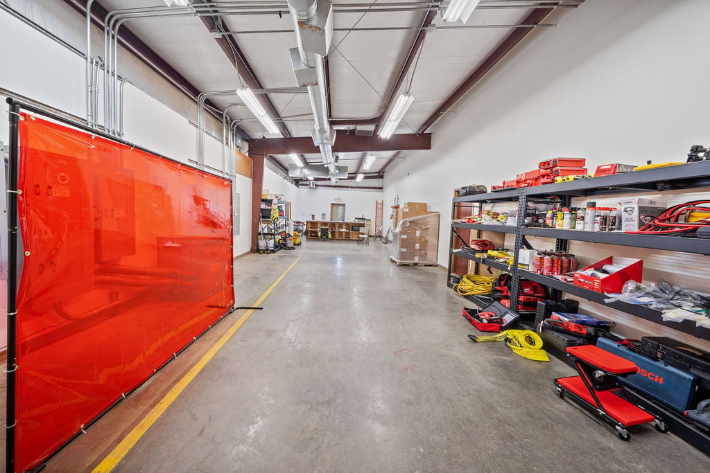 Shop interior with a red welding curtain, exposed ductwork and shelving stocked with tools and equipment.